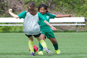 Zwei Mädchen in grünen Trikots, die auf einem grünen Rasenfeld Fußball spielen, mit einer weißen Bank, Bäumen und Pflanzen im Hintergrund und einer Wand unten.