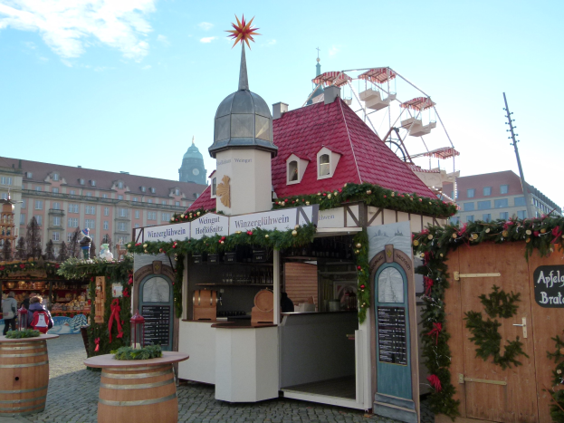 Kleines Gebäude mit rotem Dach und einem Riesenrad davor, umgeben von Festdekorationen und Menschen, mit Gebäuden, Bäumen und einem bewölkten Himmel im Hintergrund bei einem Oktoberfest-Event in München, Deutschland.