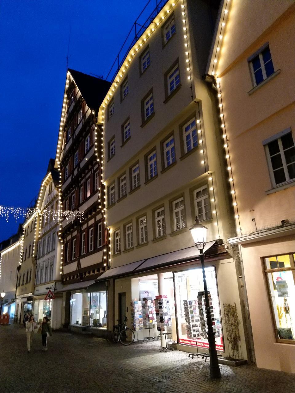 Eine festlich geschmückte Kopfsteinpflasterstraße in Rothenburg ob der Tauber, Deutschland, gesäumt von Gebäuden mit Fenstern, beleuchtet von Weihnachtslichtern mit Menschen, die vorbeigehen, und einem Fahrrad in der Nähe unter einem sichtbaren Himmel.