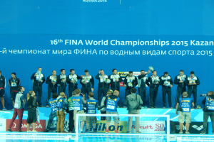 Eine Gruppe von Menschen auf dem Dach eines Schwimmbads, einige halten Kameras, mit einem Netz im Vordergrund und einem Banner mit der Aufschrift "FINA World Championships 2015 Kazan" im Hintergrund.
